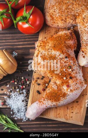 Rohe Hähnchenbeine auf einem Schneidebrett auf einem Holztisch. Mariniertes Hähnchenbein mit Gewürzen und Tomaten. Vertikales Foto Stockfoto