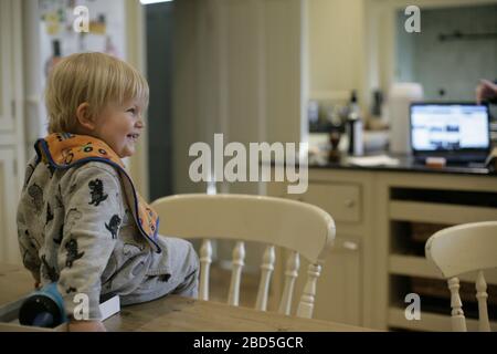 Das Kind sitzt zu Hause auf dem Tisch, während die Mutter während der Zeit der Selbstisolierung während der COVID-19-Pandemie 2020 in der Küche arbeitet Stockfoto