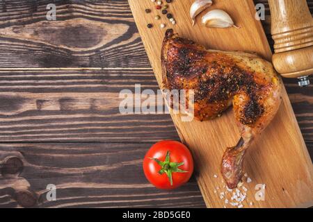 Geräuchertes Hähnchenbein auf einem Schneidebrett auf einem Holztisch. Gebratenes Hähnchenbein mit Gewürzen und Tomaten, Knoblauch und Pfeffer Stockfoto