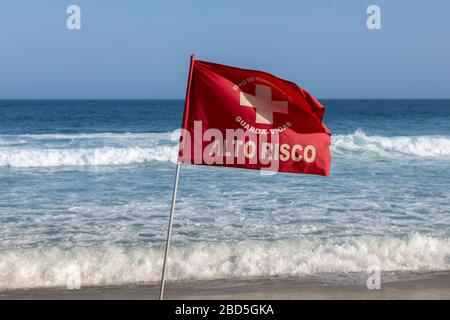 Rote Warnfahne am Strand von Leblon mit Wellen, die im Hintergrund vor einem klaren blauen Himmel aufleuchten. ÜBERSETZUNG: "FEUERWEHR. RETTUNGSSCHWIMMER. HOHES RISIKO' Stockfoto