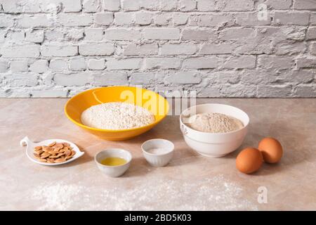 Backzutaten, Mehl, Salz, Eier und Öl in weißen und gelben Schüsseln auf Marmor-Arbeitsplatte in heller, heimeliger Küche Stockfoto