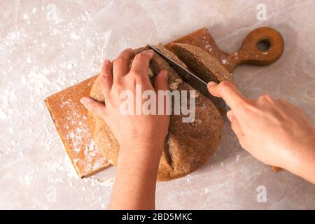 Top View Frau Hände Schneiden frisches, hausgebackenes gesundes Brot aus Roggenmehl mit Kürbiskernen auf rustikalem Holzschnittbrett auf Marmor-Arbeitsplatte Stockfoto
