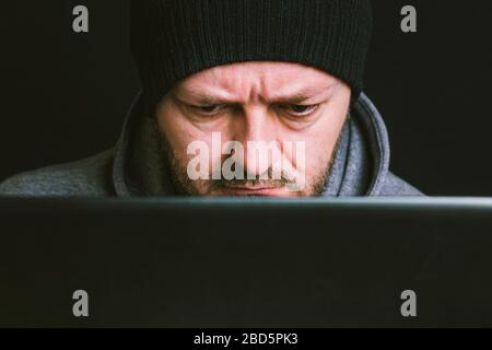 Mann trägt schwarze Mütze und Hoodie sitzt hinter dem Computer-Monitor in der Dunkelheit, Nahaufnahme, hacking Computer-System-Konzept Stockfoto