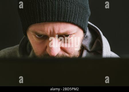 Mann trägt schwarze Mütze und Hoodie sitzt hinter dem Computer-Monitor in der Dunkelheit, Nahaufnahme, hacking Computer-System-Konzept Stockfoto