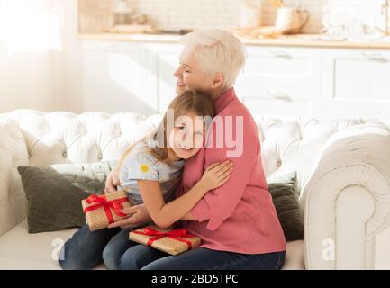 Dankbar Granny mit Geschenken, die ihr Enkelkind im Wohnzimmer umarmen Stockfoto