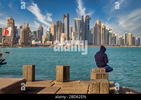 Tageslichtblick auf die Skyline von Doha Katar mit Wolken am Himmel, die Wolkenkratzer im Hintergrund zeigen und ein Mann, der auf der Daha-Cor-Nische im Vordergrund mit Katar sitzt Stockfoto