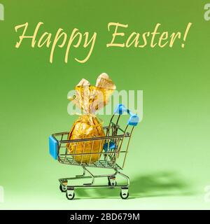 Kleiner dekorativer Supermarktkorb mit einem festlichen, in goldenem Papier gehüllten Osterei mit Aufschrift Happy Easter auf neomintem Hintergrund. Urlaub Stockfoto