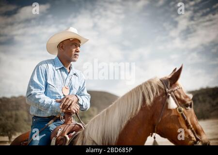 Der Mann, der einen Cowboyhut trägt, sitzt auf einem Pferd, das seine Hände auf dem Pommel ruht und blickt mit einem wachsamen Blick davon. Stockfoto