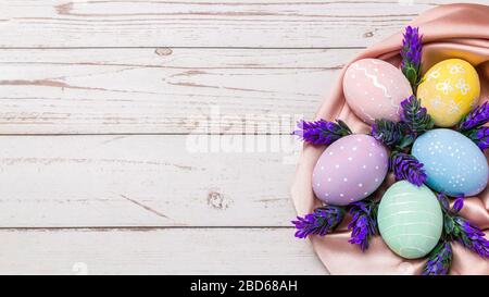 osterhintergrund mit handgemalten Pastellfarben, Eiern und frischen Lavendelblüten auf rosa Seidenstoff über hellfarbenem Holztisch Stockfoto