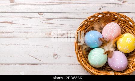 Draufsicht auf ostereier handbemalt in Pastellfarben und zarten Federn in einem Korbkorb auf Holztisch Stockfoto