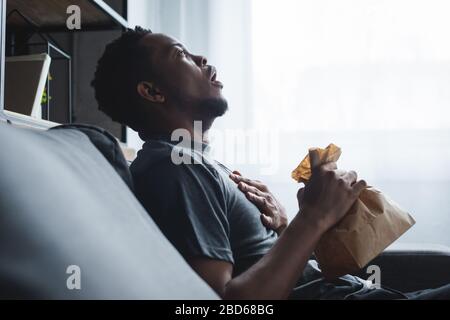 Schockierter afroamerikanischer Mann, der Papiertüte hält, während er zu Hause Panikattacken hatte Stockfoto