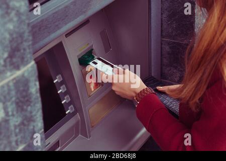 Nahaufnahme eines zugeschnittenen Bildes, in dem die Hand an einem Geldautomaten eine Debitkarte einzieht. Hand der Frau verwendet Kreditkarte, um am Bankterminal abzuheben. Moderner Stil. Geldautomat Bar t. Stockfoto