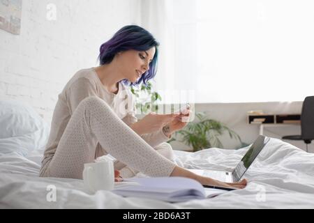 Freiberufler mit farbenfrohen Haaren mit Smartphone in der Nähe von Laptop und Tasse Tee im Bett Stockfoto