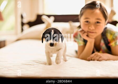 Kleine Mädchen ruht auf ihrem Kinn auf der Hand, während sie auf einem Bett mit einem Welpen liegt, der für ein Porträt posieren soll. Stockfoto