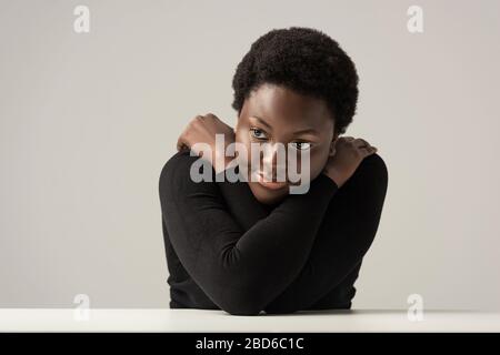 Traurige afroamerikanische Frau in schwarzem Turtleneck, die isoliert auf Grau am Tisch sitzt Stockfoto