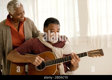 Lächelnde ältere Frau zu ihrem Ehemann spielen eine akustische Gitarre. Stockfoto