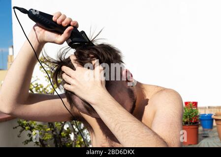 Teenager, der während der Quarantäne des Coronavirus in Barcelona, Katalonien, Spanien, mit einem Haarschneider auf dem Balkon seines Hauses seine Haare schneidet Stockfoto