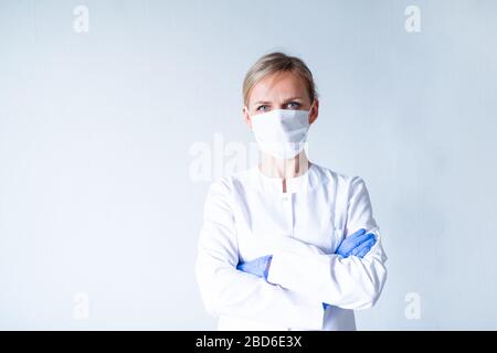 Ärztin Chirurg in weißer Uniform, blaue Schutzhandschuhe, die auf medizinische Maske aufsetzen, Hände über Grau geklemmt. Gesundheitswesen, bleiben Sie zu Hause. Copyspace Stockfoto