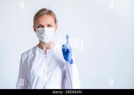 Achtung. Blonde Ärztin in weißer Uniform, blaue Schutzhandschuhe und medizinische Maske, die den Finger über Grau zeigt. Stockfoto