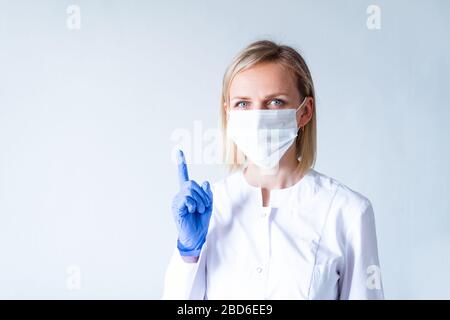 Achtung. Blonde Ärztin in weißer Uniform, blaue Schutzhandschuhe und medizinische Maske, die den Finger über Grau zeigt. Stockfoto