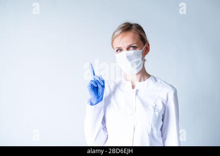 Achtung. Blonde Ärztin in weißer Uniform, blaue Schutzhandschuhe und medizinische Maske, die den Finger über Grau zeigt. Stockfoto