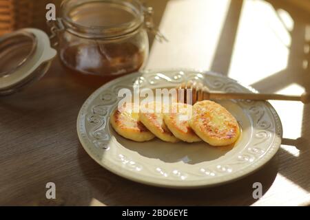 Russische und ukrainische Küche. Frühstück. Käsekuchen oder Syrniki. Käsekuchen mit Honig und einem Holzlöffel für Honig auf einem Teller auf Holz Stockfoto