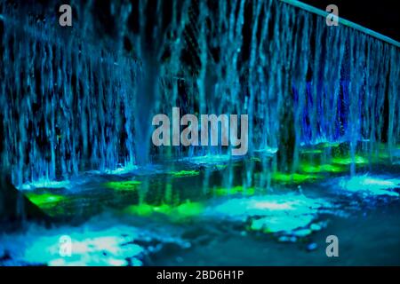 Springbrunnen mit langen Wasserstrahlen blau und grün, die in türkisfarbenes Licht übergehen. Stockfoto