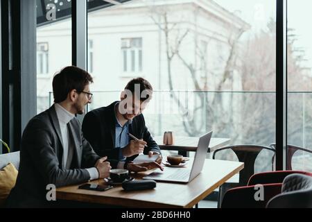 Zwei Geschäftsleute sprechen über neue Möglichkeiten, mit einem Laptop am Schreibtisch in der Nähe eines großen Fensters zu sitzen, ein Projekt zu planen, wenn sie ein Geschäftsangebot in Betracht ziehen Stockfoto