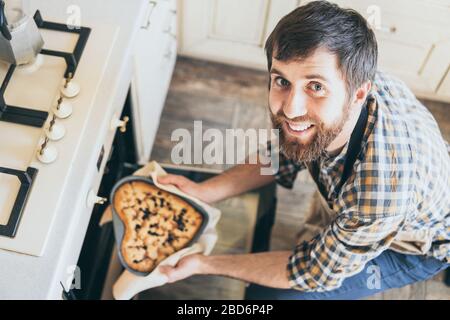 Bärtiger junger Mann, der herzförmige Beerenkuchen aus dem Ofen nimmt. Zu Hause kochen und lächeln. Stockfoto
