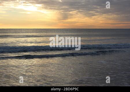 Sonnenaufgang am frühen Morgen am Strand, mit goldenen Sonnenstrahlen, die über dem Horizont aufsteigen und Wellen, die an der sandigen Küste schlagen. Stockfoto