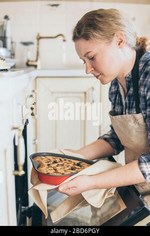 Junge blonde Frau, die herzförmige Beerenkuchen aus dem Ofen nimmt. Zu Hause kochen und lächeln. Stockfoto