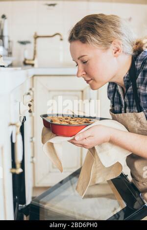 Junge blonde Frau, die herzförmige Beerenkuchen aus dem Ofen nimmt. Zu Hause kochen und mit geschlossenen Augen riechen. Stockfoto