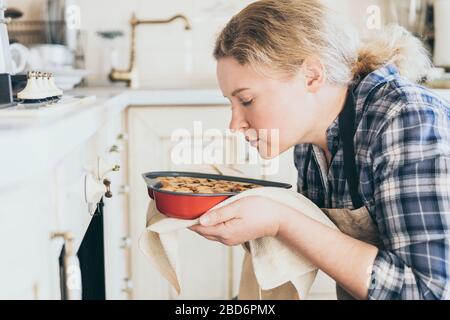 Junge blonde Frau, die herzförmige Beerenkuchen aus dem Ofen nimmt. Zu Hause kochen und mit geschlossenen Augen riechen. Stockfoto