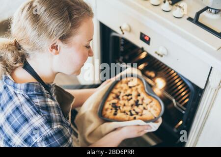 Junge blonde Frau, die herzförmige Beerenkuchen aus dem Ofen nimmt. Zu Hause kochen und lächeln. Stockfoto