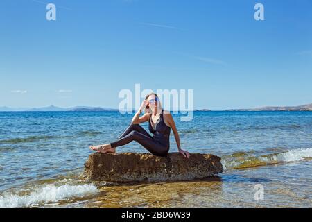 Junge Frau in Sportbekleidung sitzt auf einem Felsbrocken im Meer. Stockfoto