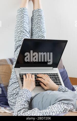 Eine Frau in geblümten Nachtwäsche liegt auf dem Rücken, ihre Beine sind gerade nach oben gerichtet und arbeiten auf ihrem Laptop. Konzept, während zu Hause zu bleiben Stockfoto