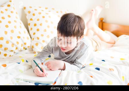 Schuljunge im Bett und Hausaufgaben zu Hause machen, in Notizbuch schreiben. Online-Fernunterricht. Quarantäne. Das Konzept, zu Hause zu bleiben Stockfoto
