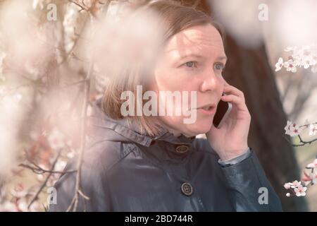 Ernsthafte Frau, die im Frühling auf dem Handy unter dem blühenden Kirschbaum spricht Stockfoto