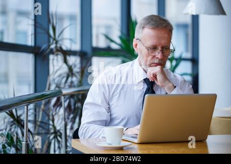 Freiberufliche Männer arbeiten in einem Café an einem neuen Geschäftsprojekt. Sitzt an einem großen Fenster am Tisch. Schaut auf einen Laptop-Bildschirm mit einer Tasse Kaffee Stockfoto