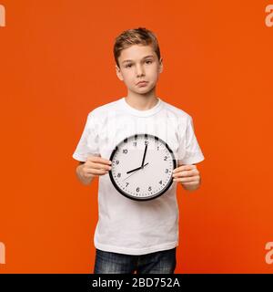 Verärgerter Teenager-Junge mit großer Uhr, orangefarbenem Studiohintergrund Stockfoto