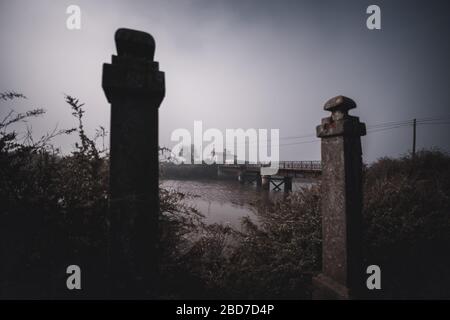 Eine weit abgewinkelt dunkle Moody Landschaft mit verfallenen Steinpfosten als Tore in einem Unschärfe-Vordergrund und einer verlassenen rostigen Brücke, die zu verlassenen Häusern führt Stockfoto