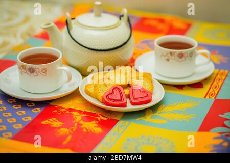 Zeit für eine Teepause. 2 Tassen Heißgetränk, leckere Plätzchen, herzförmige Süßigkeiten, Pralinen auf einem Tisch. Datum, Freunde besuchen, großes Mutterhaus, Brea Stockfoto