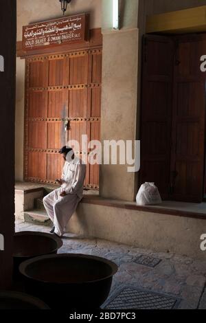 Ein omanischer Mann in Geschirrspüler sitzt mit seinem Handy neben verzierten und dekorativen traditionellen Türen im alten Souk von Nizwa im Oman Stockfoto