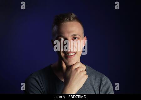 Junge kaukasische blonde lächelnde Mann in grauem T-Shirt auf dunkelviolettem Hintergrund berührt sein Kinn Stockfoto