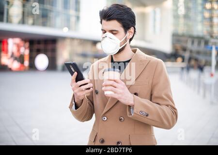 Maskierter junger Mann, der sein Handy benutzt und eine Tasse Kaffee hält, während er in einer Stadt, einem Coronavirus und einem Pandemiekonzept spaziert Stockfoto