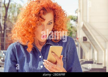 Nahaufnahme des Porträts schockierter Frauen mit roten lockigen Haaren, beim betrachten des Mobiltelefons, das einige schlechte Nachrichten oder Fotos mit verblüfften Emotionen im Gesicht sieht. Ist Stockfoto