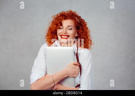 Ich liebe meinen Laptop. Rote, lockige Geschäftsfrau in weißem Hemd umarmend Computer lächelnd fröhliche Augen geschlossen isoliert auf grauer Hintergrundwand. Stockfoto