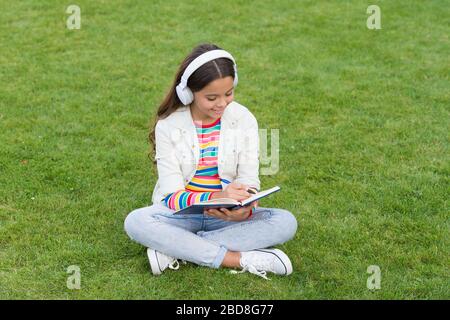 Liedtexte schreiben. Fröhliches Kinderkonzept. Kind schreibt Erinnerungen in Tagebuch. Schulmädchen entspannen sich mit Notizbuch. Glückliches Kind hört Musik in Kopfhörern. Kleines Mädchen auf grünem Gras. Kind genießt Lied in der Natur im Park. Stockfoto