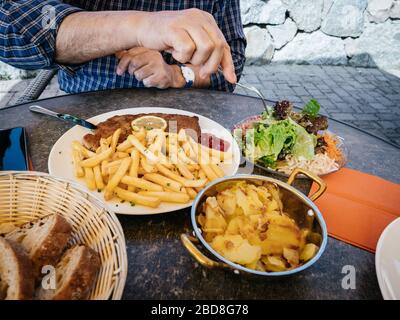 Detail der männlichen Chefhand, die mit gebackenen Pommes frites und Pommes frites aus großem Gericht deutschen Schnitzel isst - Salat schmeckt Stockfoto