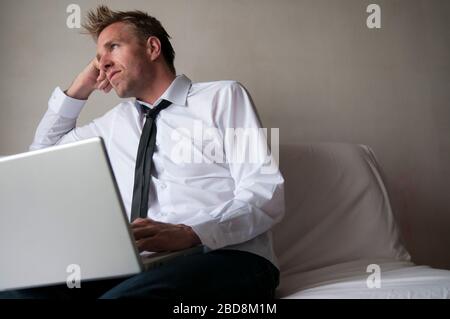 Gelangweilter Mann in eleganten lässigen Jeans sitzt auf dem Sofa Surfen seinen Laptop-Computer Stockfoto